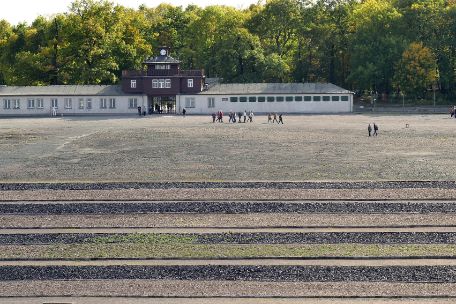 Zu sehen ist der ehemalige Appellplatz als eine große Freifläche vor dem Lagertor, welches im Hintergrund zu sehen ist. Im Vordergrung des Bildes sind auf dem Boden die Umrisse der ehemaligen Baracken des Lagers durch Felder aus Schlackesteinen sichtbar gemacht, die sich gegenüber dem übrigen Kies dunkel abheben..