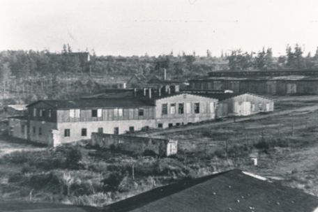 Das Bild zeigt das ehemalige Werkstattgelände nach Auflösung des Speziallagers. Im Zentrum des Bildes ist ein langes, mehrgeschossiges Gebäude zu sehen. Im Hintergrund eine pferdestallartige Baracke. Der Komplex ist von ausgedünnten Baumreihen umgeben. 