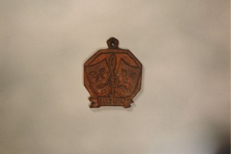 Holzanhänger aus einer lagerinternen Produktion von Goethes Faust von 1946. Der Anhänger ist achteckig und zeigt eine lachende und eine traurige Maske nebeneinander.
