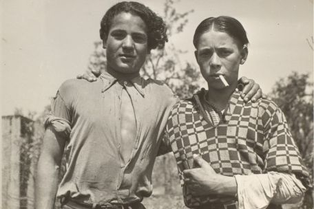 Two boys clasp each other by the shoulders and look into the camera. One has a cigarette in his mouth. 