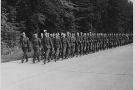 SS-Angehörige einer Rekruteneinheit der SS-Totenkopfstandarte 14 beim Rückmarsch nach ihrer Vereidigung auf der Blutstraße. In Dreierreihen geht eine lange Schlange von Uniformierten die aus Betonplatten bestehende Straße durch den Wald entlang. 