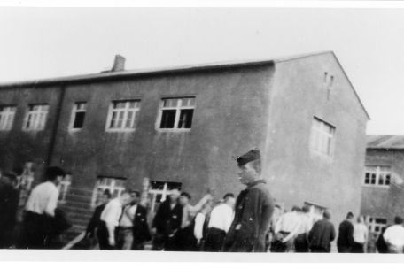 Häftlinge laufen vor einem der Steinblocks von links nach rechts. Die Mütze des im Vordergrund stehenden Häftlings ist retuschiert.