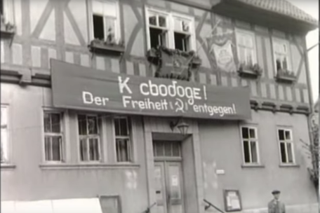 A screenshot from a movie. A half-timbered house is decorated with a banner showing a hammer and sickle, Cyrillic lettering and the words "Towards Freedom". 