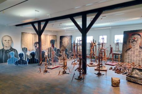 View of the installation Remineszenzen by Josef Szajna in the art exhibition of the Buchenwald Memorial. Arid, abstract wooden frames fill the space, vaguely reminiscent of people. In the background, larger-than-life portraits, a large list, and shoes on the floor. 