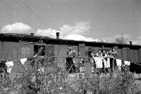 In front of a wooden barrack, eight people stand leaning against a railing on which laundry has been hung out to dry. Almost all of them smile friendly into the camera.