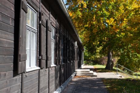 The photo shows the black wooden exterior of the reconstructed wooden barracks. On the right you can see that it was built on a slope. A staircase leads down. Next to the stairs is a tree whose leaves have turned autumnal.
