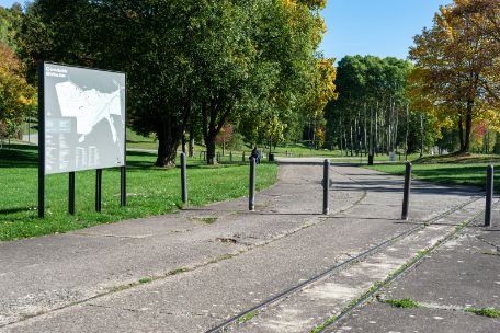 Ein festmontierter Geländeplan, der am Wegrand steht. Im Hintergrund herbstliche Bäume. Im Vordergrund sind auf dem Boden Schienen für einen Lorenwagen zu erkennen.