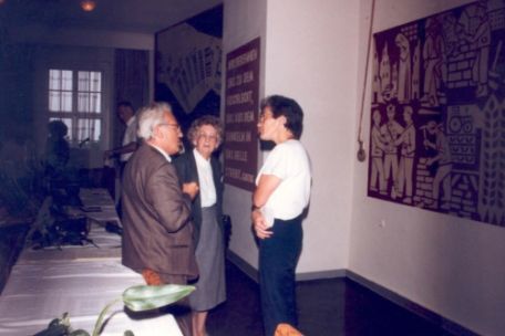 Drei Menschen stehen in einem Raum und diskutieren. Die beiden links und in der Mitte stehenden Personen sind älter. Im Raum befinden sich ein langer Tisch, ein Fenster und zwei Wandbilder.