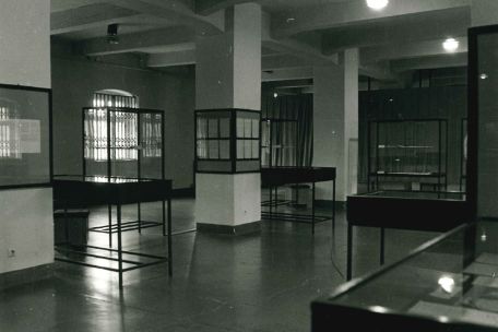 Auf einer Schwarz-Weiß-Aufnahme ist ein Raum mit mehreren Säulen zu sehen. Die Fenster im Hintergrund sind vergittert. An den Wänden befinden sich Schaukästen mit Dokumenten. Im Raum stehen mehrere Tisch und Schrankvitrinen.