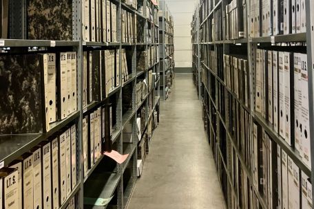 Akten stehen in Ordnern in langezogenen Regalen auf vier Regalböden verteilt. Die Regale erstrecken sich auf die gesamte Länge des Raumes.
