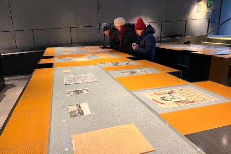Drei Personen stehen an einem sehr großen orangefarbenen Ausstellungstisch. Auf grauen Flächen sind Bilder und Schriftstücke zu sehen. Der Tisch erstreckt sich über die ganze Länge des Raumes. Hinter den Personen steht ein weiterer baugleicher Ausstellungstisch.