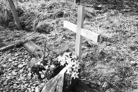 Holzstämme und Steine markieren ein Grab im Wald. Auf dem Grab steht ein Holzkreuz. Vor dem Kreuz liegen ein Kranz und Blumen. Es ist ein Schwarz-Weiß-Bild.
