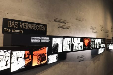 An einer unverputzten Betonwand hängen schwarz gerahmte Lichtkästen mit alten Fotos. Die Fotos sehen aus wie Scherenschnitte. An der Betonwand stehe Zitate in Deutsch und Englisch. Auf schwarzem Untergrund steht die weiße Überschrift "Das Verbrechen". Darunter steht kleiner geschrieben "The Atrocity"