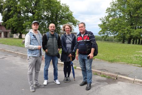 Auf dem Bild sind vier Personen zu kernnen, die auf einer asphaltierten Straße stehen. Im Hintergrund sind Teile des ehemaligen Lagertors erkennbar. Die Personen im Vordergrund stehen nebeneinander und schauen in die Kamera. Von links nach rechts sind zuerst zwei Männer zu sehen. Neben ihnen steht eine Frau und rechts von ihr ein weiterer Mann.