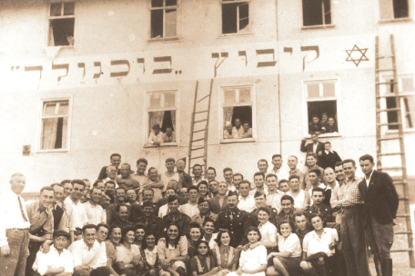 Historisches Gruppenfoto vor einem Gebäude: Zahlreiche junge Menschen stehen und sitzen dicht gedrängt zusammen und blicken in die Kamera. An der Fassade über ihnen befindet sich eine hebräische Inschrift sowie ein Davidstern; einige Personen schauen aus geöffneten Fenstern, und Leitern lehnen an der Hauswand.