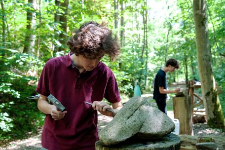 Two teenage volunteers work on memorial stones of the Buchenwald Railway Memorial Trail with hammer and chisel