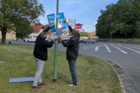 Zwei Männer entfernen ein Plakat der AfD von einer Laterne am Parkplatz der Gedenkstätte. Es trägt des Slogan "Mut zur Wahrheit"