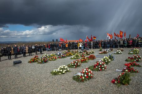 Wreaths are laid in rows on the roll call square. They are surrounded by a barrier tape. Behind them are numerous people, some of whom are carrying flags. Dark clouds provide dramatic lighting. 
