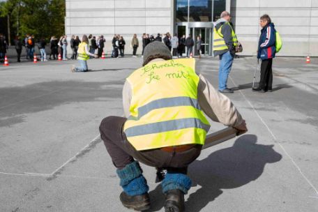 A man kneels on a stone square and wears a yellow high-visibility vest with the inscription "Write with" on his back. In his hands he holds a sticky board, which he puts straight in front of him. In the background, other people with high-visibility vests are standing and kneeling in the square.