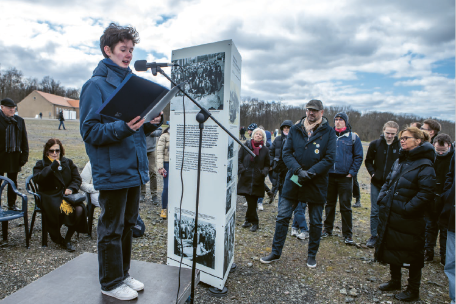 Ein junger Mensch steht draußen auf einer kleinen Bühne und liest aus einem Ordner in ein Mikrofon. Um ihn herum hat sich eine Gruppe von Zuhörer:innen versammelt, die aufmerksam zuhören. Im Hintergrund steht eine Informationstafel mit historischen Fotos und Texten, die auf einen Gedenk- oder Erinnerungsort hinweist.