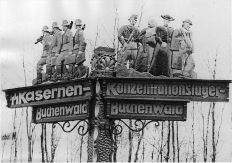  Ein handgeschnitzter Wegweiser zu den SS-Kasernen Buchenwald und dem KZ Buchenwald am Carachoweg. Über den Schriftzügen sind aus Holz Geschnitzte Figuren zu erkennen. Links: SS-Soldaten, rechts: Ein SS-Mann der drei Personen vor sich her treibt. Die Figuren sind als Mönch, Priester, und Mann mit Anzug und Hut, dargestellt.