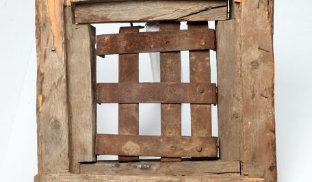 A wooden door made of rough beams. A window in it is barred with wide iron bars. 