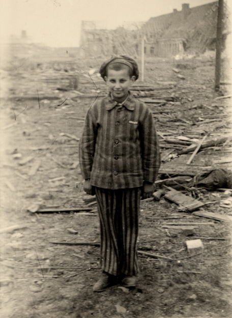 A boy in prisoner's clothing stands in front of rubble and smiles at the camera.