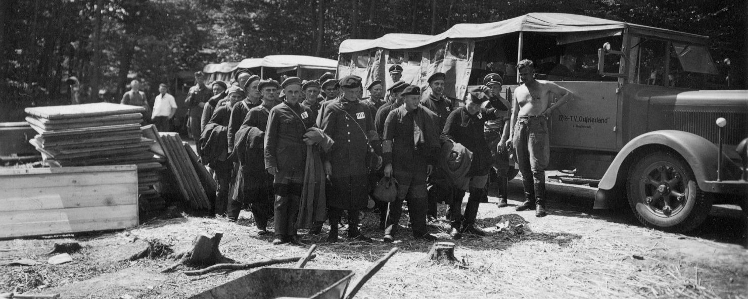In Viererreihe aufgestellte Häftlinge stehen inmitten hoher Bäume. Neben ihnen liegen Baustoffe und eine Schubkarre. Im Hintergrund stehen SS-Wachmänner vor ihren Mannschaftswagen.