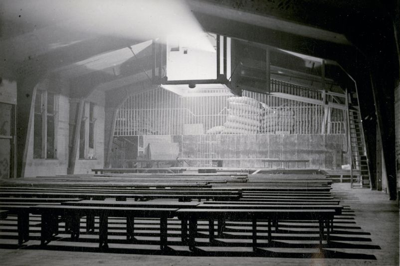 You can see an empty barrack with benches in the centre. The picture was taken from the screen. The benches are facing the screen. A projector hangs from the ceiling in a large box from which a cone of light shines forwards.