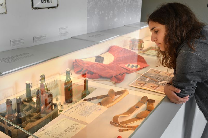 A visitor leans over a display case and looks inside. Inside are bottles, leather goods, documents and a large swastika flag. 