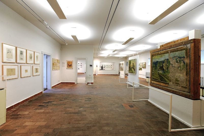 Das Bild zeigt einen Blick in einen der Ausstellungsräume der Kunstausstellung, Links an der Wand sind mehrere kleinere Zeichnungen in Bilderrahmen an der wand zu sehen. Rechts sind in der Mitte des Raumes drei aus der ursprünglichen Wand geschnittene Reliefgemälde zu sehen.