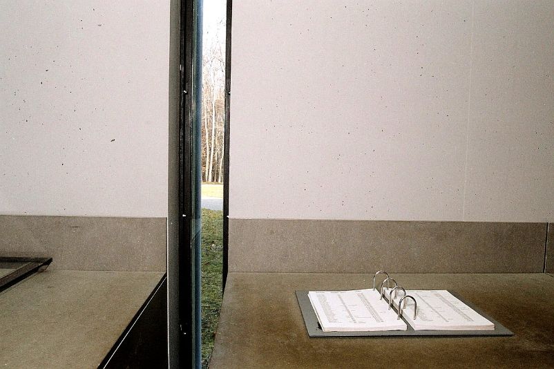 Ein verglaster Sehschlitz neben einem aufgeschlagenen Totenbuch auf einem Wandtisch ermöglicht den Blick von der Ausstellung nach draußen auf das Gräberfeld.
