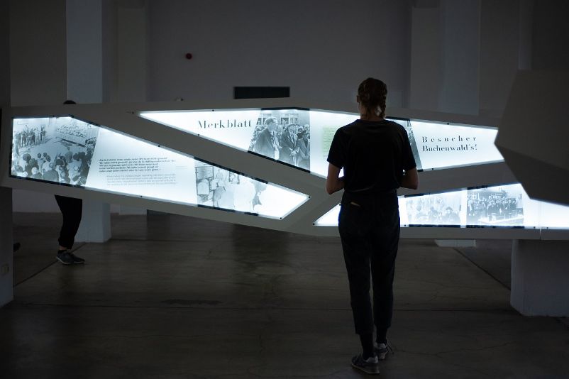 A young visitor stands in front of illuminated exhibition panels. The panels are arranged asymmetrically and stretch across the room at chest level along a beam-shaped construct.