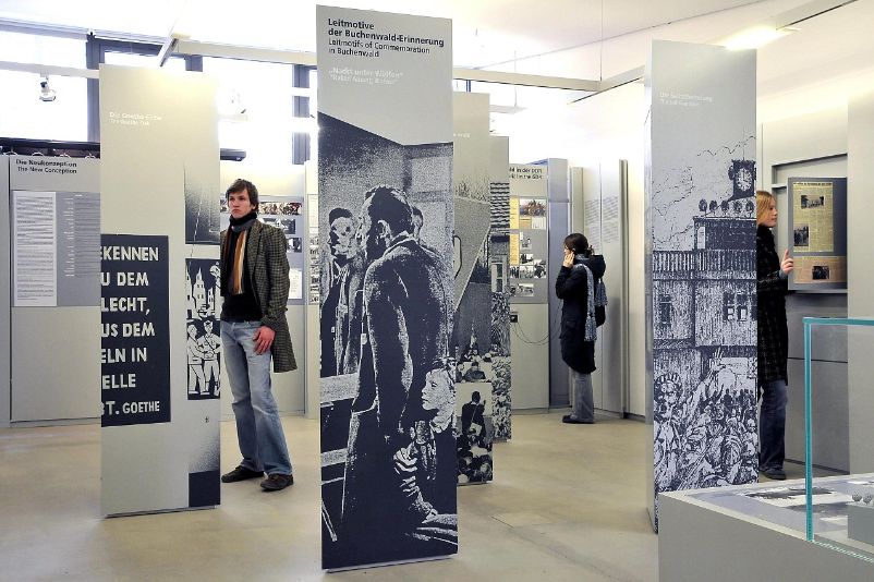 Das Foto zeigt das Ausstellungsgebäude von innen. Große, nahezu bis zur Decke gehende Informationswände stehen in der Mitte des Raumes, dazwischen befinden sich vereinzelt lesende Besucher:innen.