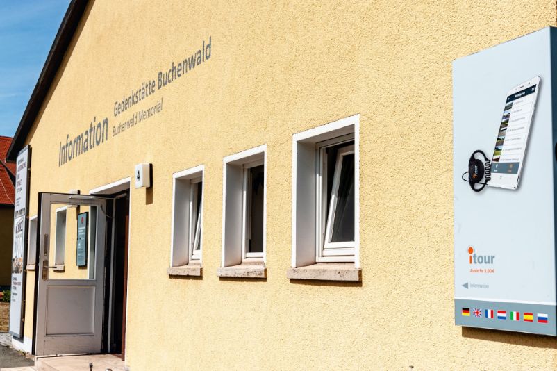 Der Eingang der Besuchsinformation. Über der offen stehenden Tür weist ein Schriftzug auf die Besuchsinformation im Inneren hin. An der Hausecke rechts im Bild weist ein Schild auf die Möglichkeit zum Ausleihen eines Audioguides hin.