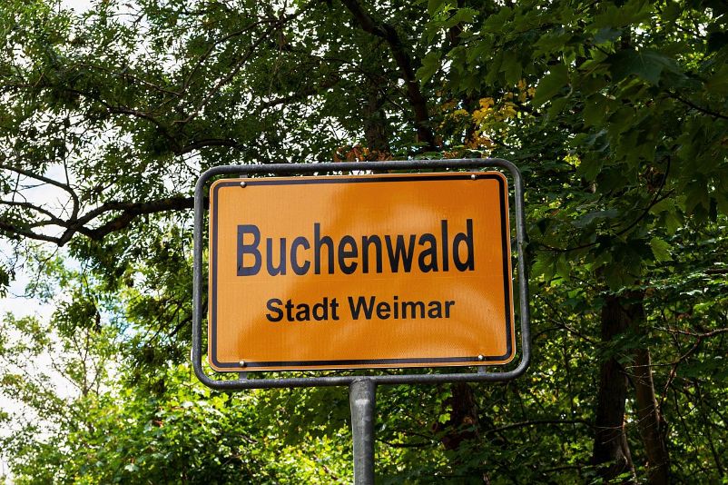 The yellow place-name sign "Buchenwald Stadt Weimar". Behind it trees.