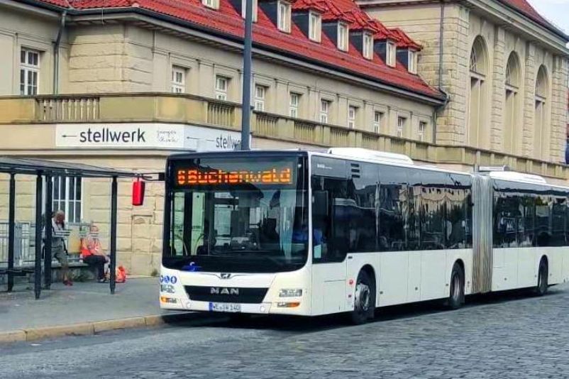 Ein weißer Bus der Linie 6 nach Buchenwald fährt die Bushaltestelle vor dem im Hintergrund sichtbaren Weimarer Hauptbahnhof an.