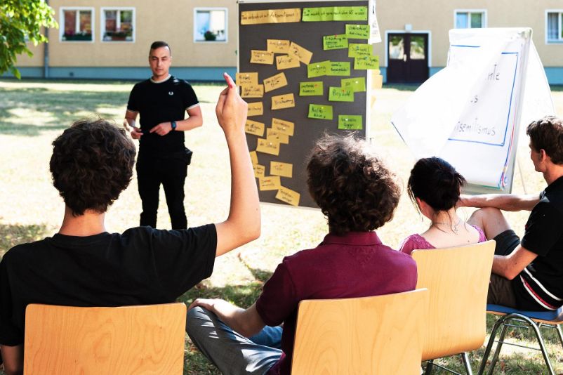 Vier Jugendliche sitzen auf Stühlen auf einer wiese und sind einem jungen Referenten zugewandt. Einer der Jugendlichen hebt den Arm, um sich zu melden. Neben dem Referenten ist ein Flipchart mit der Aufschrift "Antisemitismus erkennen"