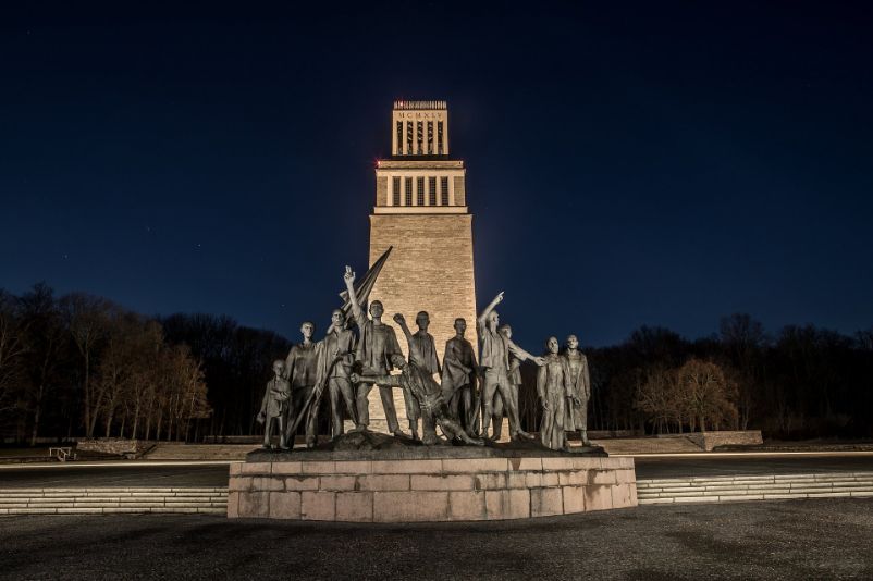 The group of figures at the Buchenwald Memorial in front of the memorial's bell tower at night. Both the group of figures and the bell tower are illuminated.