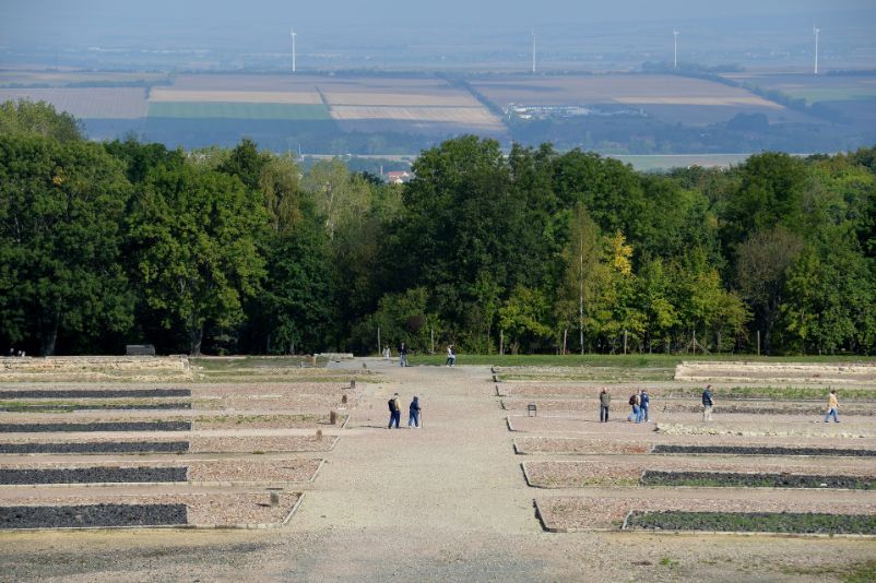 Blick auf eine der Lagerstraßen. Rechts und Links sind die ehemaligen Lagerblöcke duch dunkle Schlackesteinfelder sichtbar gemacht. In der Ferne geht der Blick ins Tal. 