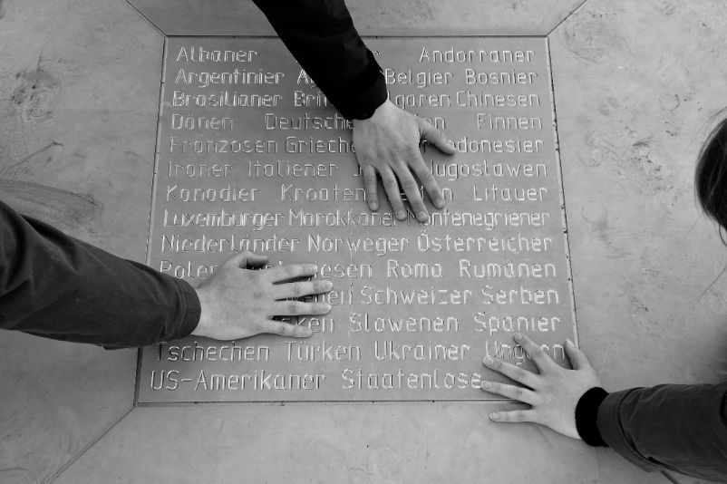 Drei Hände von unterschiedlichen Personen berühren die beheizte Mittelplatte des Gedenkzeichen auf dem Appellplatz. Auf der Platte sind Nationalitäten zu lesen