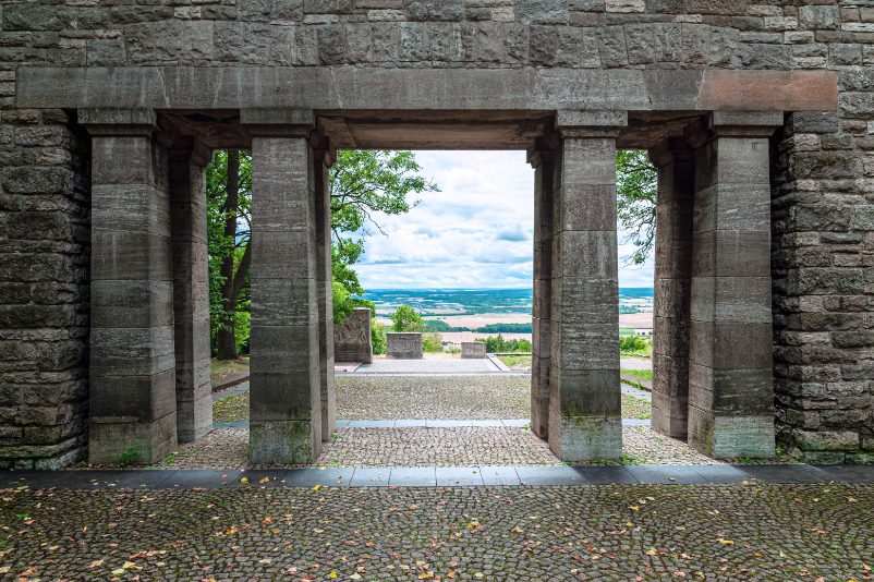Blick durch den Torbogen, der den Eingang der Mahnmalsanlage markiert. Dahinter führt der Weg bergab, entlang an massiven Stelen. 