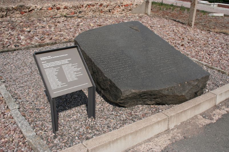 Ein dunkelgrauer Gedenkstein mit einer Inschrift in kyrillischer Schrift. Daneben ein Schild mit einer Übersetzung. 
