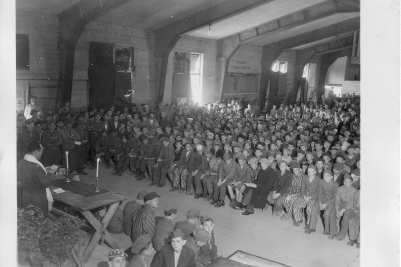 In einer großen Halle sitzen Angehörige der US Army und befreite Häftlinge. Am linken Bildrand steht ein Mann und hält einen jüdischen Gottesdienst ab.