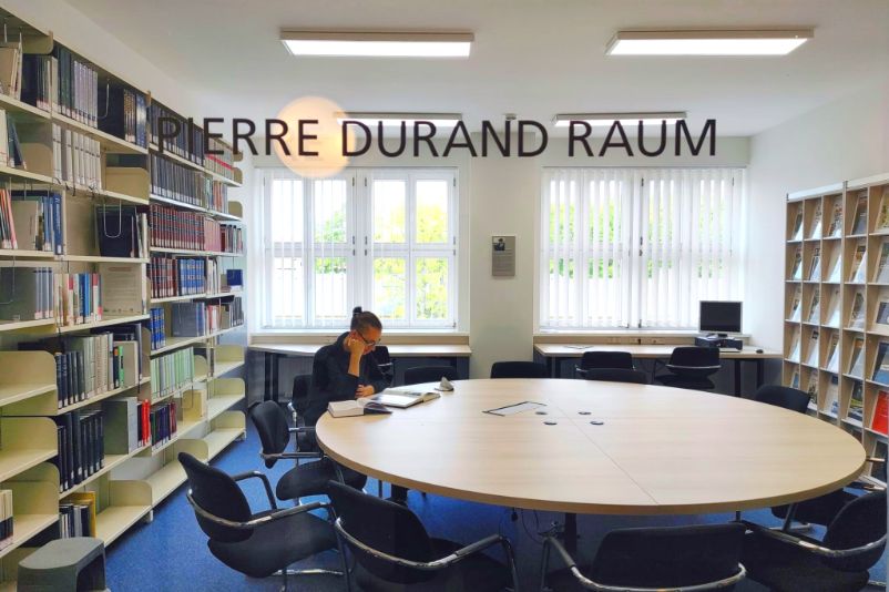 Ein Nutzer sitzt am Runden Holztisch in der Mitte des Lesesaals der Gedenkstättenbibliothek und ließt. Das Foto ist vom Flur aus durch die gläserne Tennwand fotografiert an der der Schriftzug "Pierre Durand Raum" zu erkennen ist.