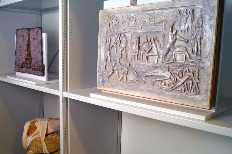 Three works of art on a shelf. The upper two are bronze plates and show abstracted scenes from the camp with violence and the dead. The lower one consists of a stack of stones held together by a ribbon. 