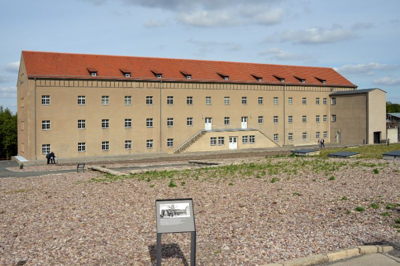 Der Blick über das Gelände aus auf das ehemalige Kammergebäude von außen. Im Vordergrund steht ein Informationsschild. Es zeigt Informationen zum Gebäude und ein altes Foto. 