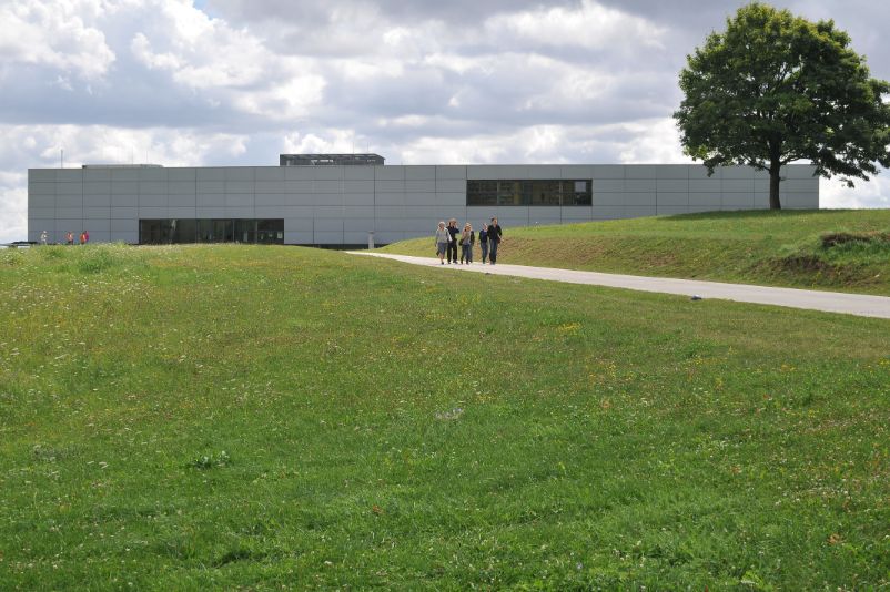 Frontal view of the new building of the memorial. It is a modern, multi-story, gray low-rise building. A group of young visitors is moving along the access path away from the building in the direction of the former camp grounds.