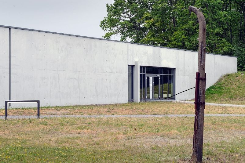 Im Zentrum ist das graue Ausstellungsgebäude zu sehen. Vor dem Ausstellungsgebäude ist Wiese, rechts im Hintergrund ein großer Baum zu sehen. Im Vordergrund steht ein Pfeiler des ehemaligen Lagerzauns.