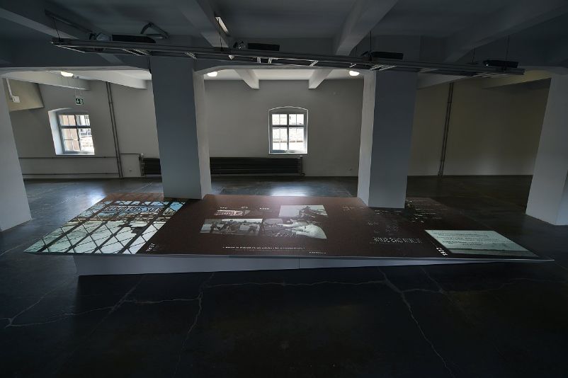 Erdgeschoss der Ausstellung. Am Beginn der KZ-Ausstellung steht im Erdgeschoss eine leicht schräge Platte, auf die eine audio-visuelle Videocollage projiziert wird. Sonst ist der Raum leer. 